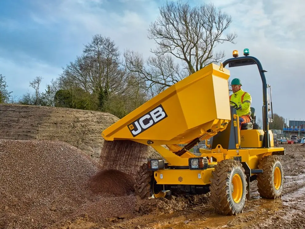 Hiretech A construction worker operates a yellow JCB dump truck, unloading gravel onto a pile at a muddy worksite with bare trees in the background. Hire2buy