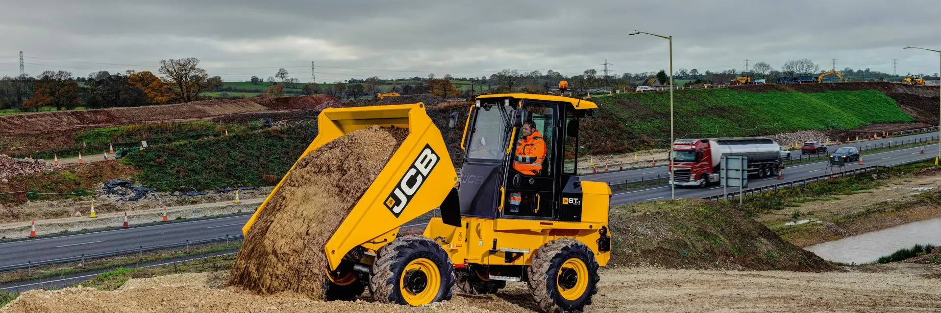 Hiretech A worker operates a yellow JCB dump truck, unloading sand or soil at a construction site next to a busy multi-lane road, with grassy hills and cloudy skies in the background. Hire2buy
