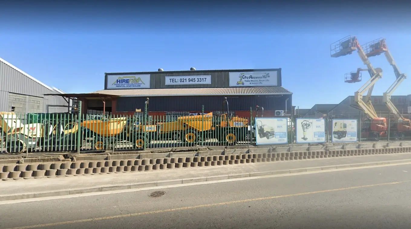 Hiretech A construction equipment rental facility with various yellow and orange machinery behind a green fence, including dumpers and aerial lifts. A large sign with contact details is visible on the building. Hire2buy